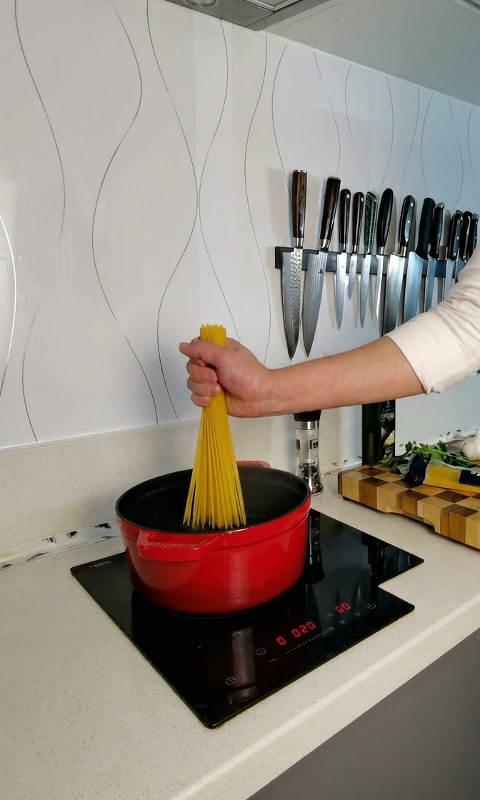 A hand holding a bundle of dry spaghetti over a red pot of boiling water, twisting the strands to fan them out.