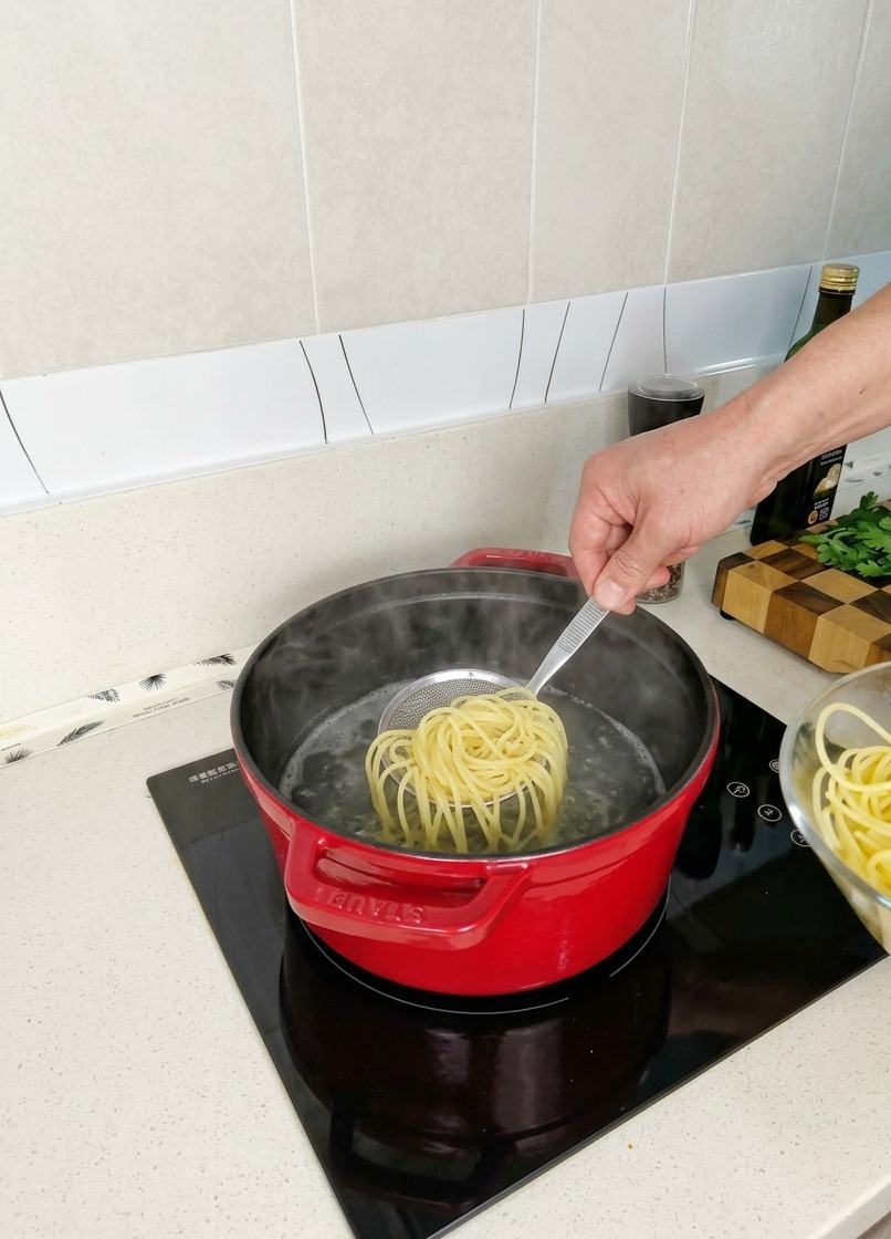 Lifting a nest of partially cooked spaghetti from a red pot of steaming water using a small mesh strainer.
