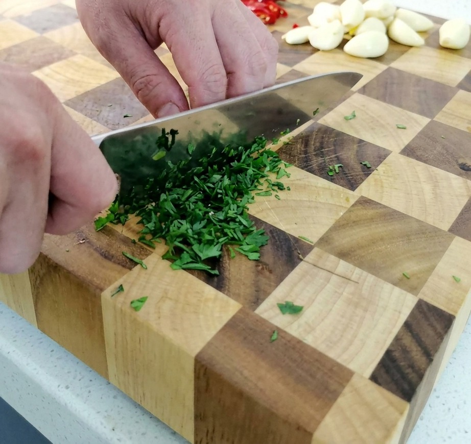 A persons hands using a chefs knife to finely mince a bunch of fresh green parsley on a wooden checkerboard cutting board.