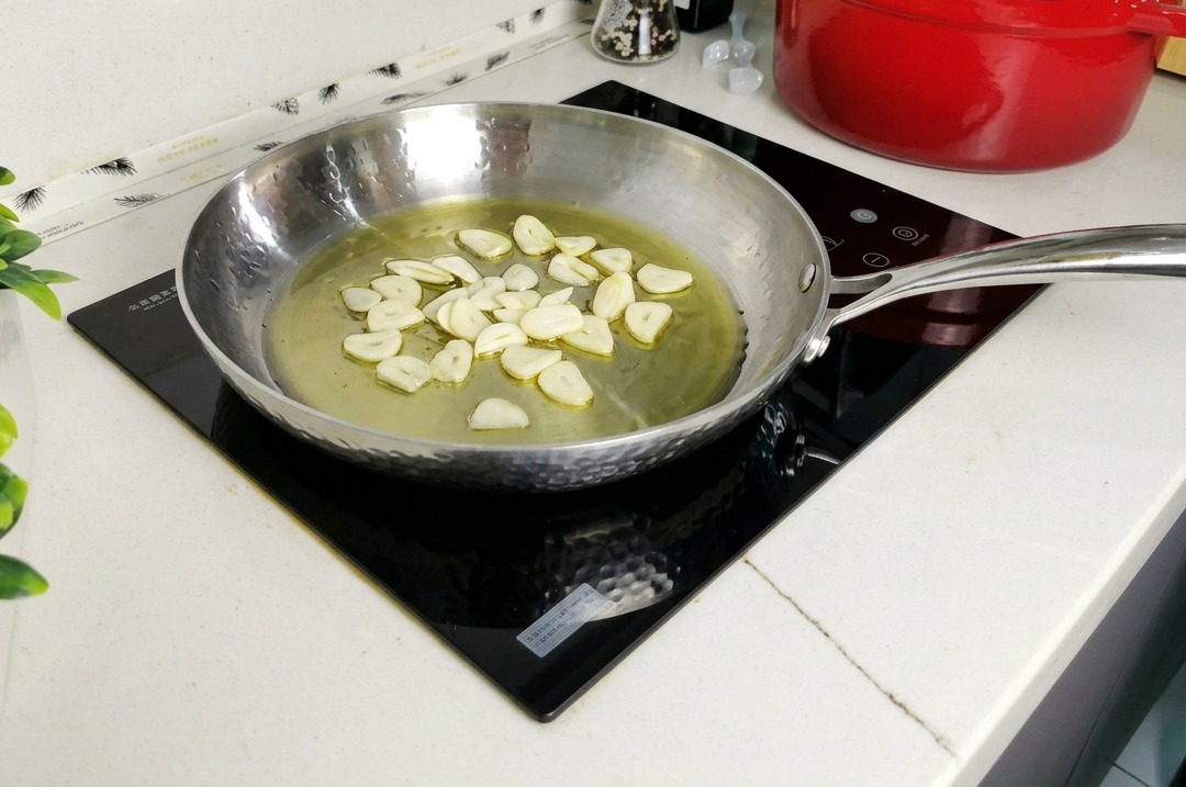 Thin slices of garlic and green parsley stems simmering in a pool of olive oil inside a hammered stainless steel frying pan.