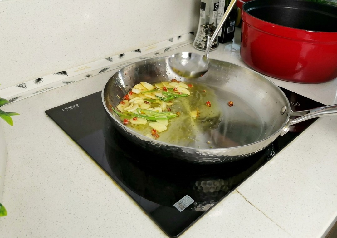 A ladle pouring cloudy pasta water into a stainless steel skillet containing sautéed garlic slices, chilies, and parsley stems.