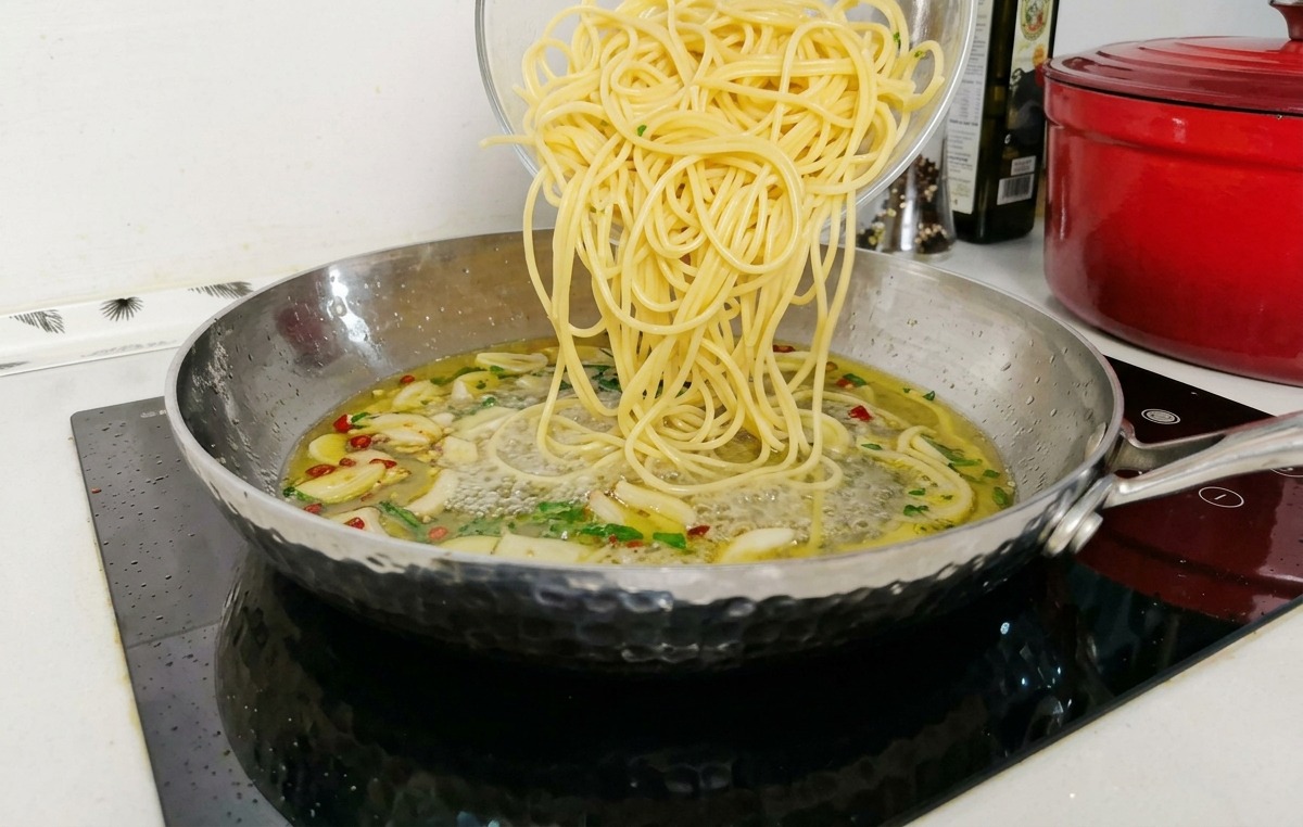 Cooked spaghetti being poured from a glass bowl into a stainless steel pan filled with a simmering garlic and oil sauce base.