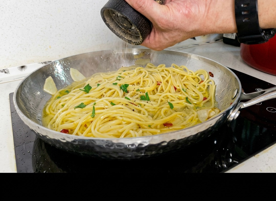 A chef grinding fresh black pepper from a mill over a pan of spaghetti pasta tossed with garlic, chili, and parsley.