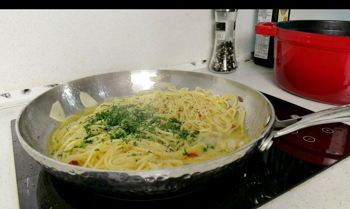 A stainless steel pan filled with spaghetti in a light sauce, topped with a pile of freshly minced green parsley.
