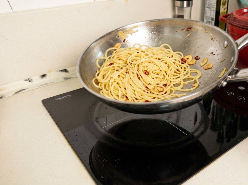 A stainless steel pan being tossed in mid-air, with spaghetti noodles being coated in a glossy, emulsified sauce.