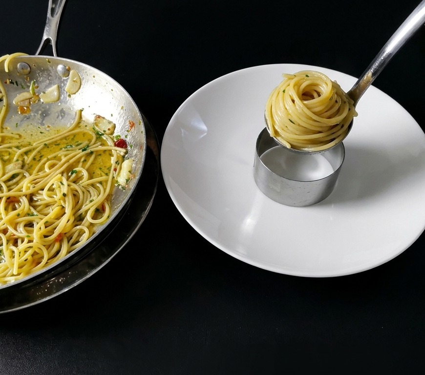 A close-up shot of spaghetti being twirled into a neat cylinder inside a metal ring mold on a clean white plate using culinary tweezers and a ladle.