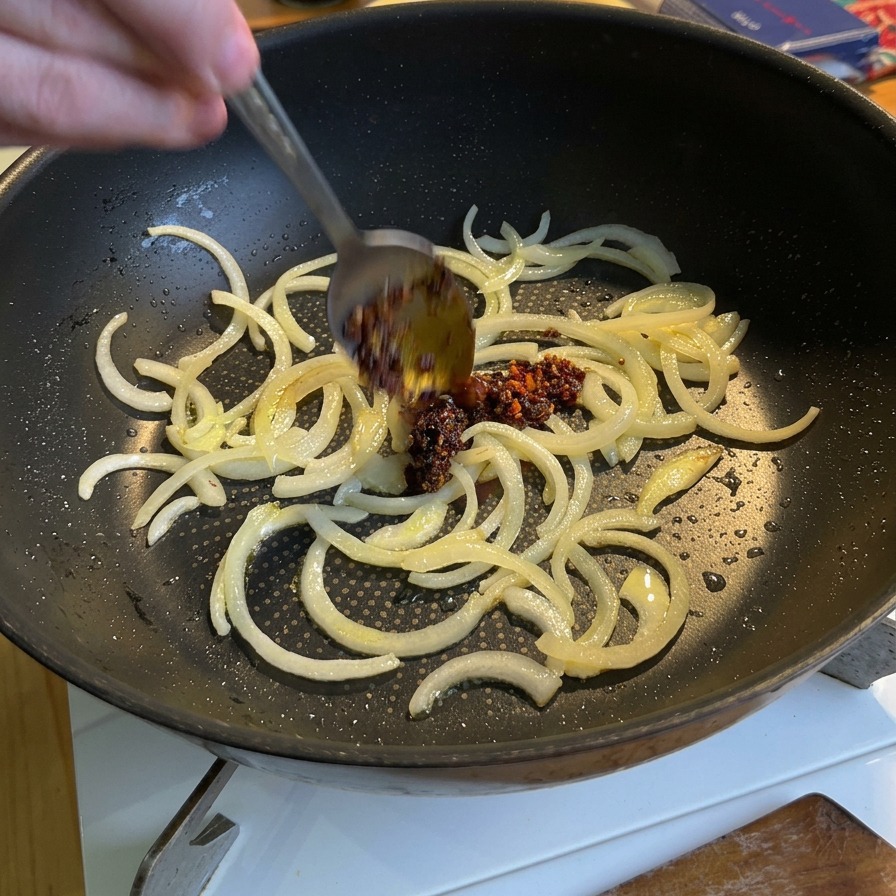 A spoon adding vibrant red chili oil into a pan containing partially cooked sliced white onions.