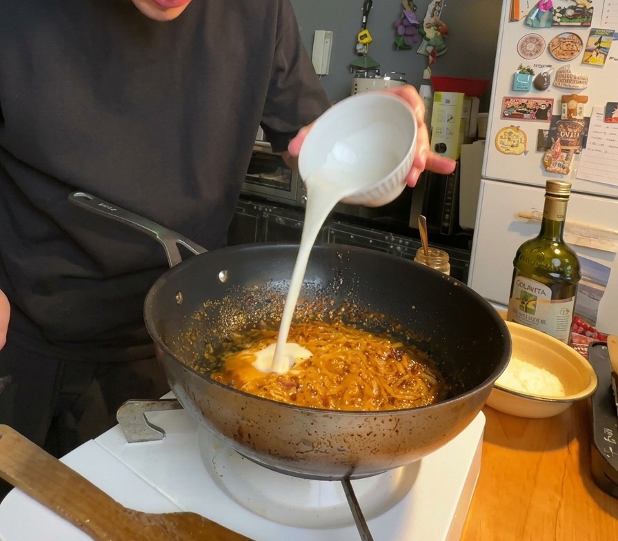 Heavy cream being poured into a pan of reddish, simmering chili and onion sauce.