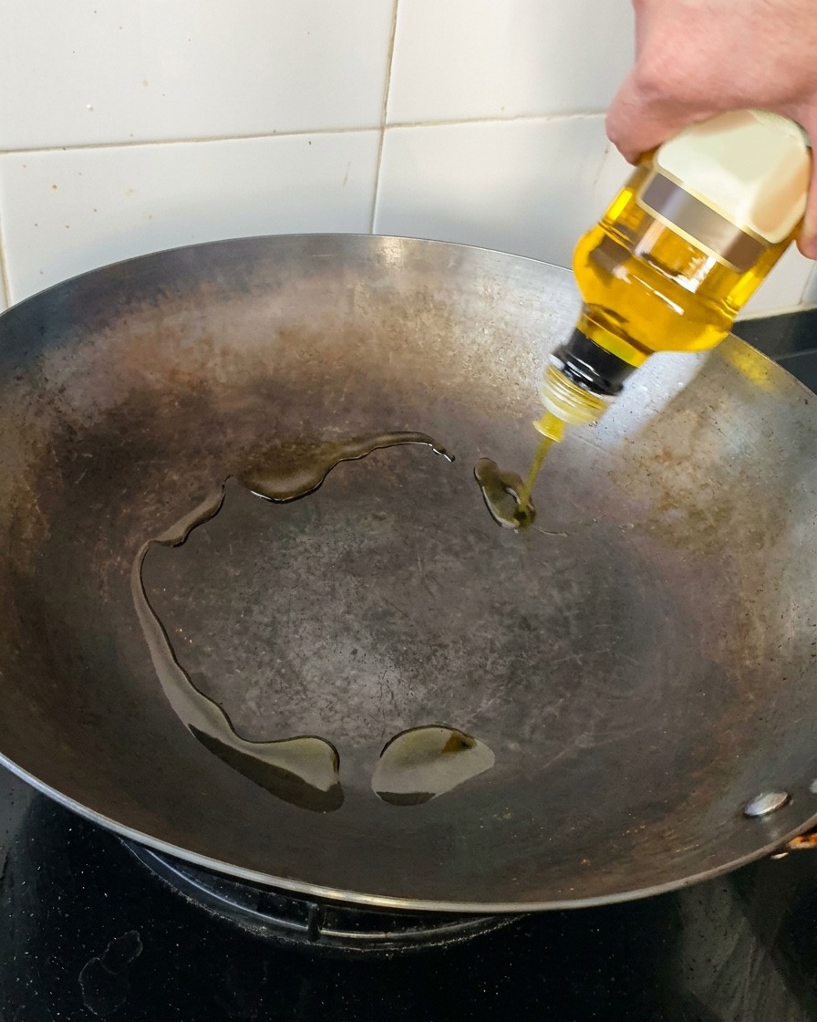 A hand pouring golden olive oil from a clear glass bottle into a dark, seasoned carbon steel wok.