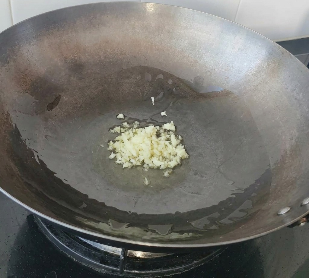 Minced garlic sizzling in a pool of olive oil at the bottom of a large carbon steel wok.