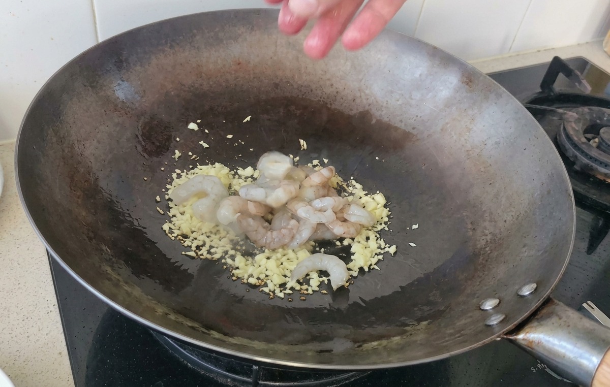 A handful of raw, translucent shrimp being dropped into a hot wok containing sautéed garlic.