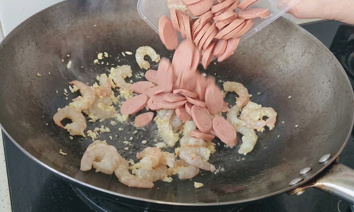 Sliced sausages being poured from a plastic container into a wok filled with stir-frying shrimp and garlic.