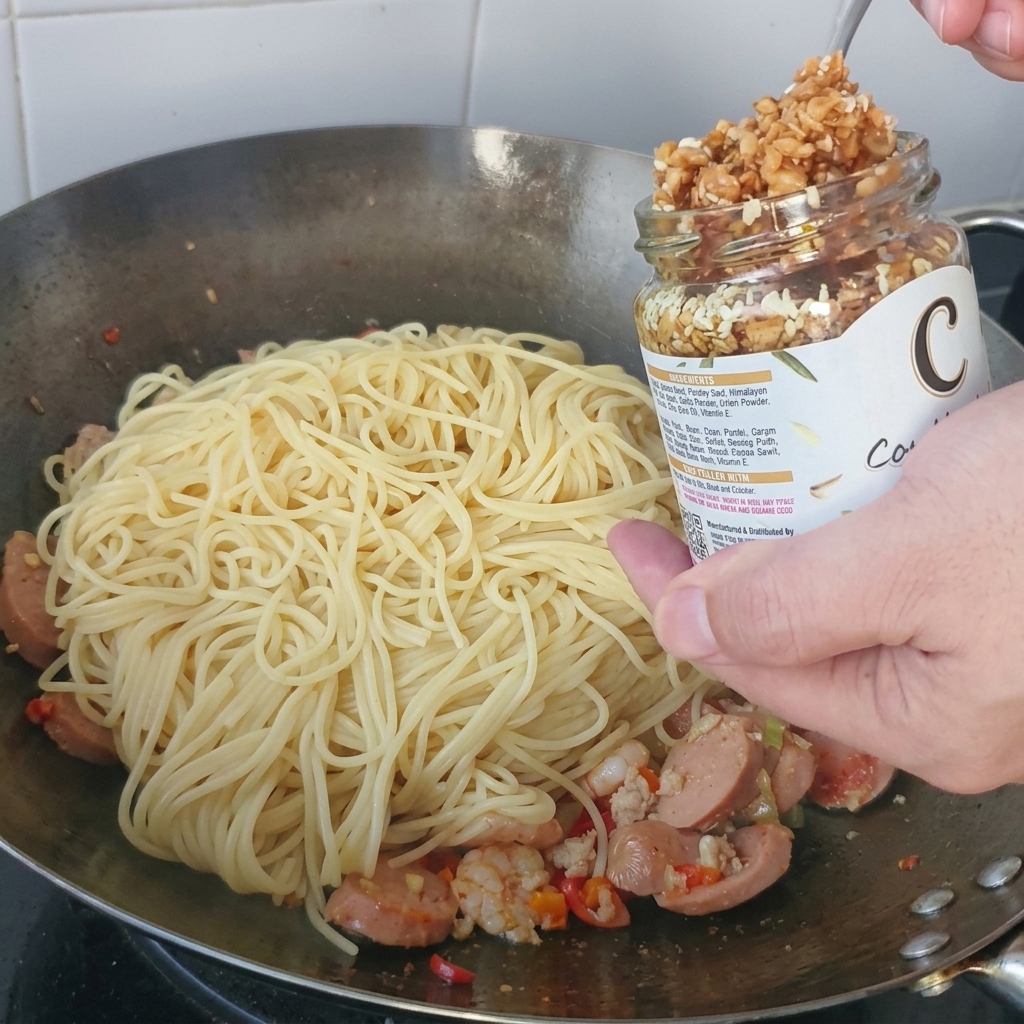 Crispy golden garlic being spooned from a glass jar onto a mound of spaghetti in a metal wok.