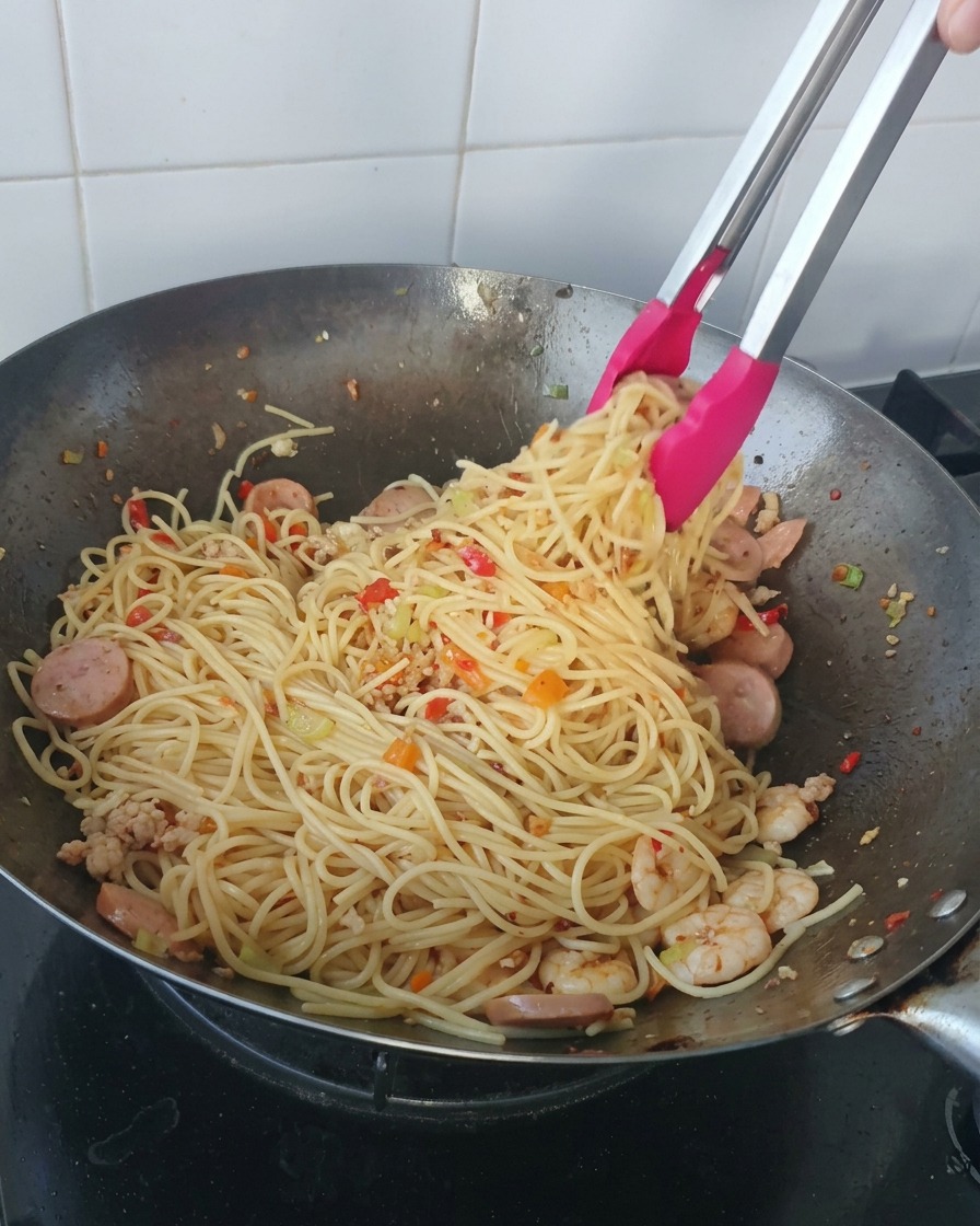 Pink-tipped tongs lifting a large portion of spaghetti mixed with shrimp and sliced sausages from a wok.