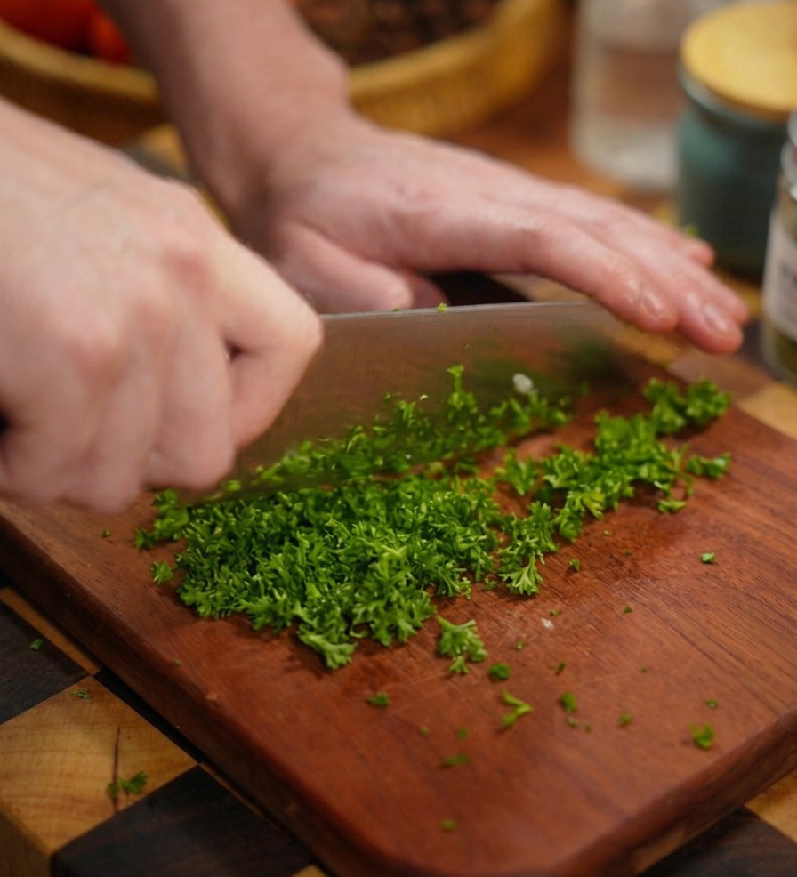 A close-up shot of hands using a large knife to finely chop bright green parsley on a rich brown wooden cutting board.
