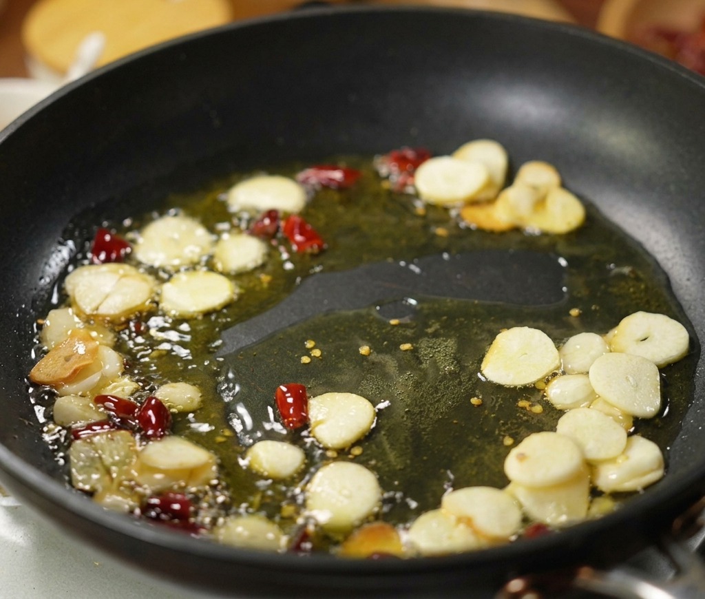 Thin slices of garlic and pieces of dried red chili peppers sizzling in a shallow pool of olive oil in a black frying pan.