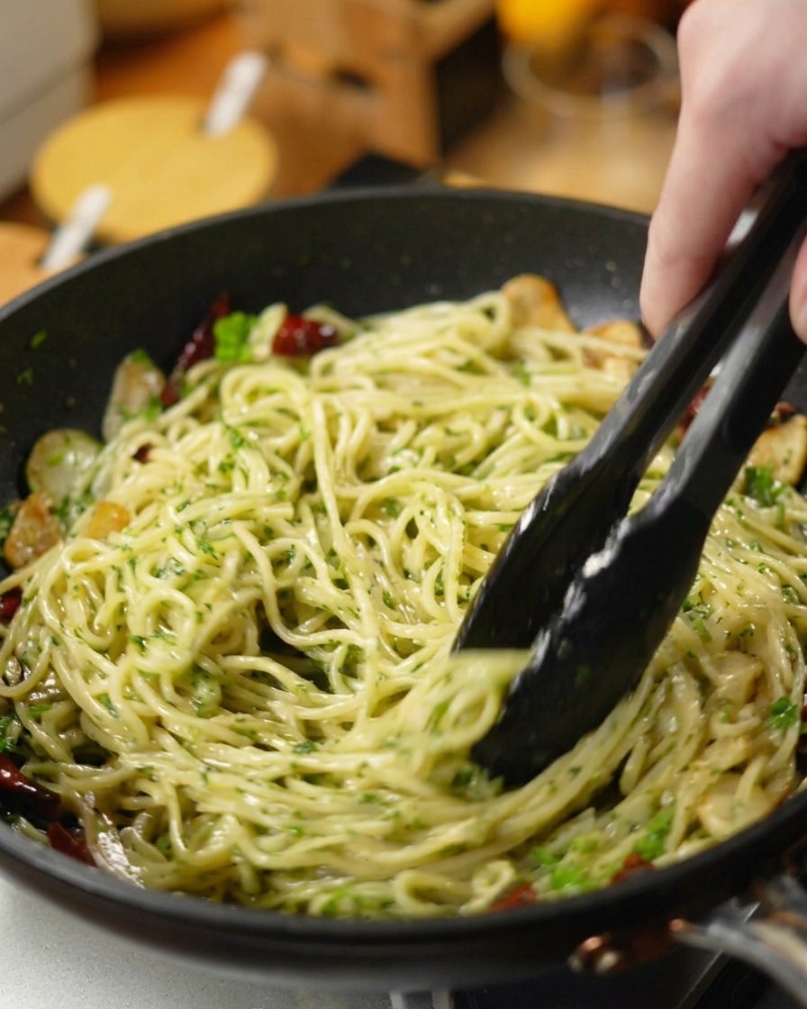 Black tongs tossing spaghetti, chopped parsley, garlic, and chilies in a dark skillet.