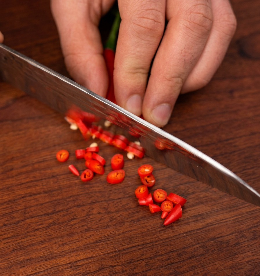 A sharp chefs knife slicing small red birds eye chilies into thin rounds on a dark wooden cutting board.