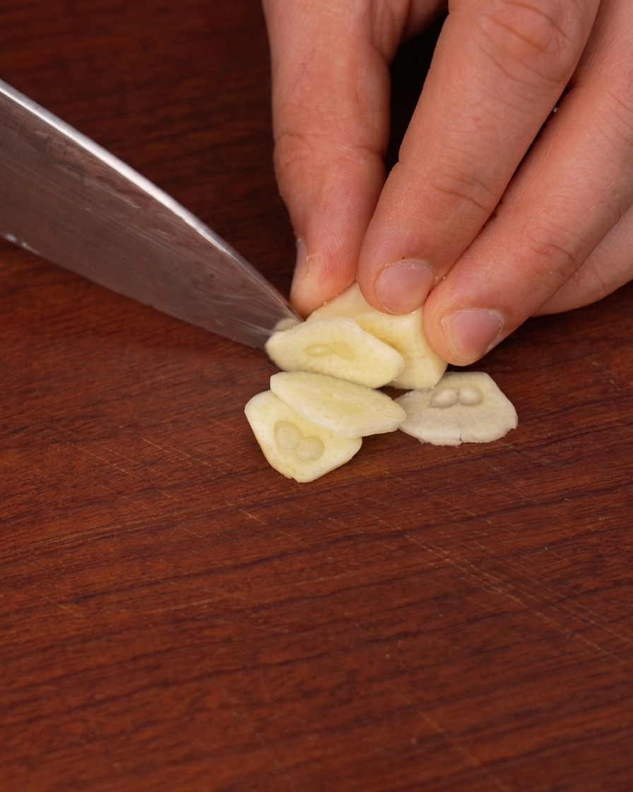 Thin, translucent slices of garlic being cut from a whole clove on a wooden surface.