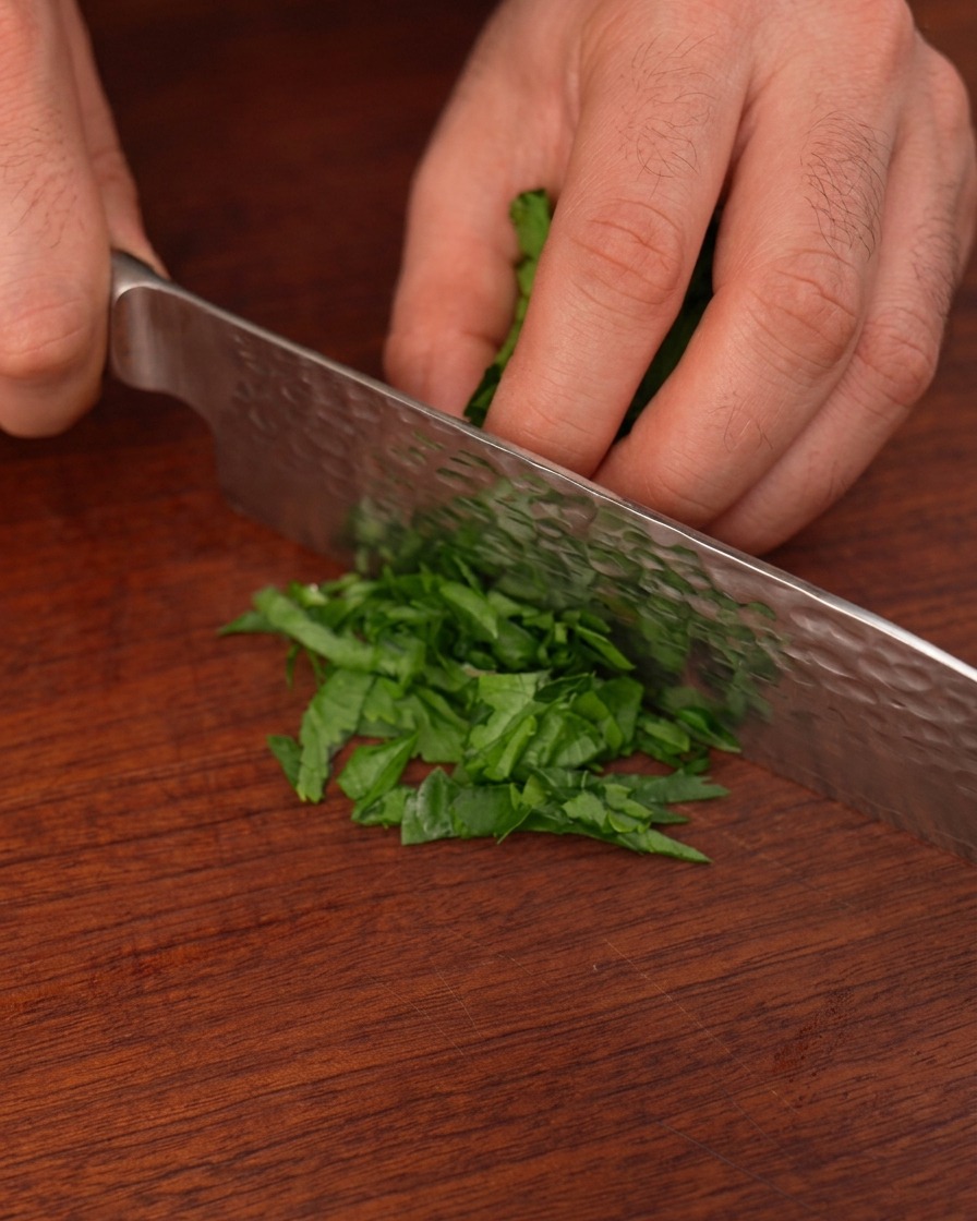 Fresh green parsley leaves being finely minced with a hammered steel chefs knife on a wood board.