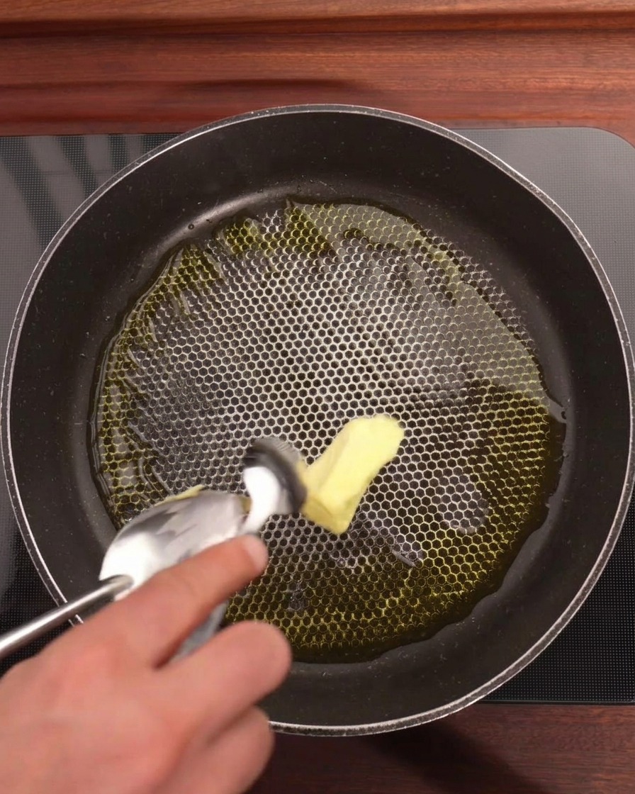 A hand using a spoon to add a knob of yellow butter into a honeycomb-textured pan containing olive oil, sliced garlic, and red chili rings.
