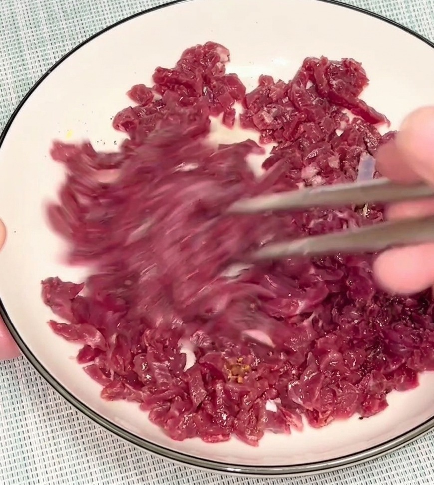 A person using silver chopsticks to mix pieces of raw, red beef in a white bowl to distribute the marinade.