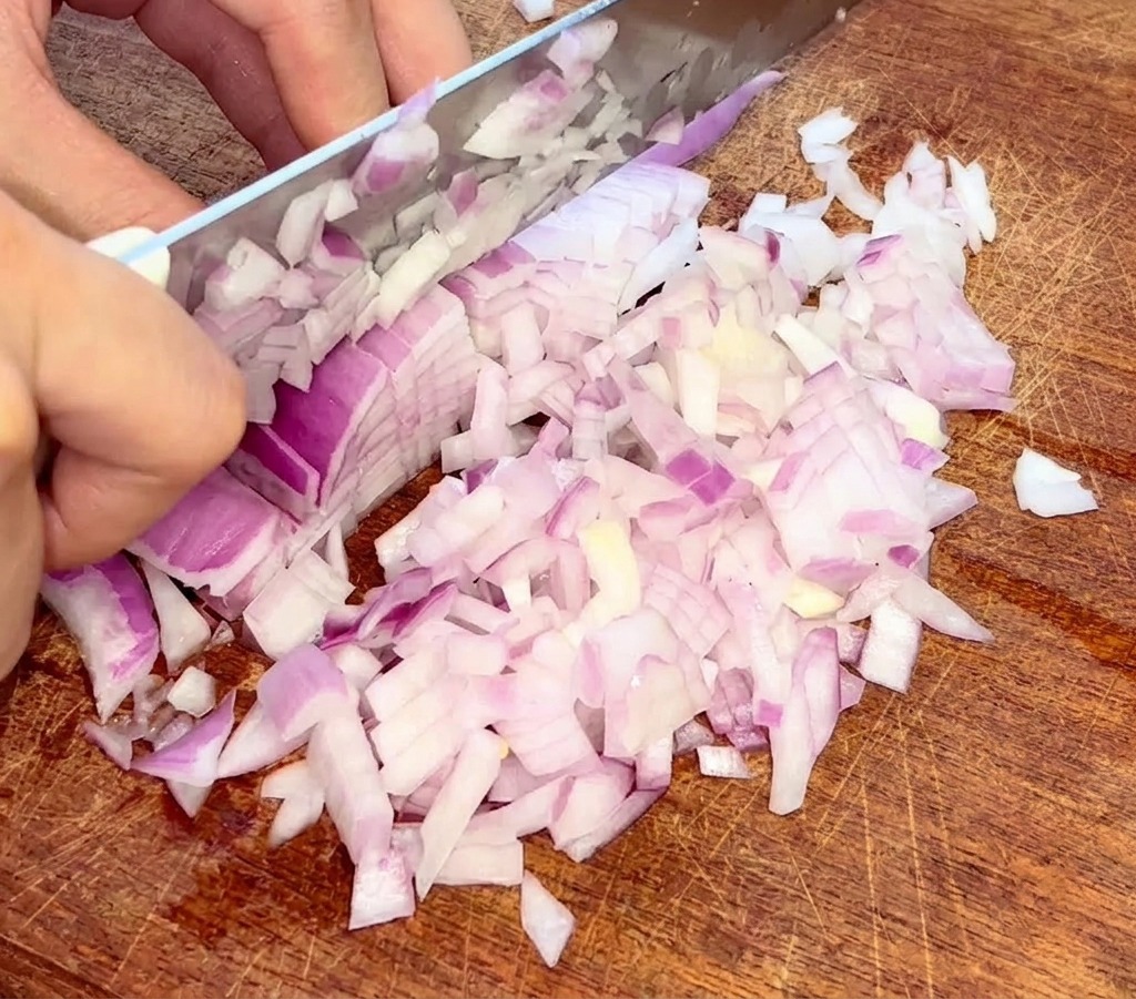 A close-up shot of hands using a knife to finely dice a red onion on a wooden cutting board.