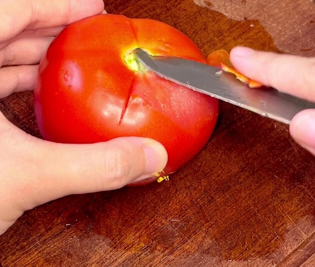Hands carefully using a paring knife to peel the skin off a whole blanched tomato.