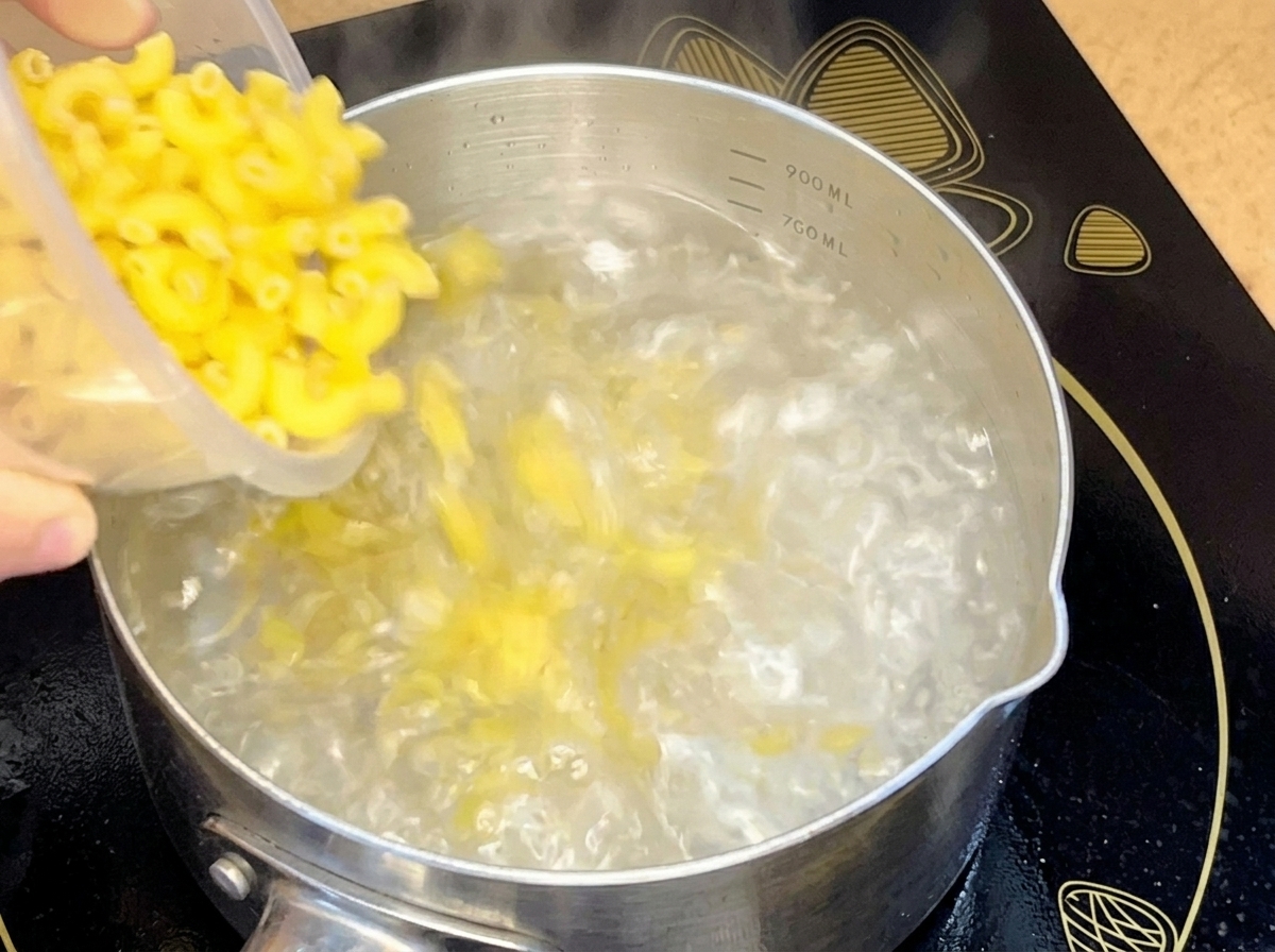 Dry elbow macaroni being poured from a container into a stainless steel pot of boiling water.