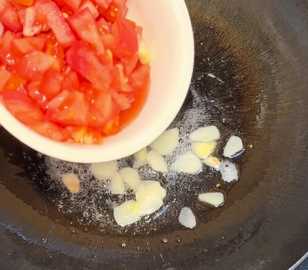 Diced tomatoes being poured from a white bowl into a pan with hot oil and golden sautéed garlic slices.