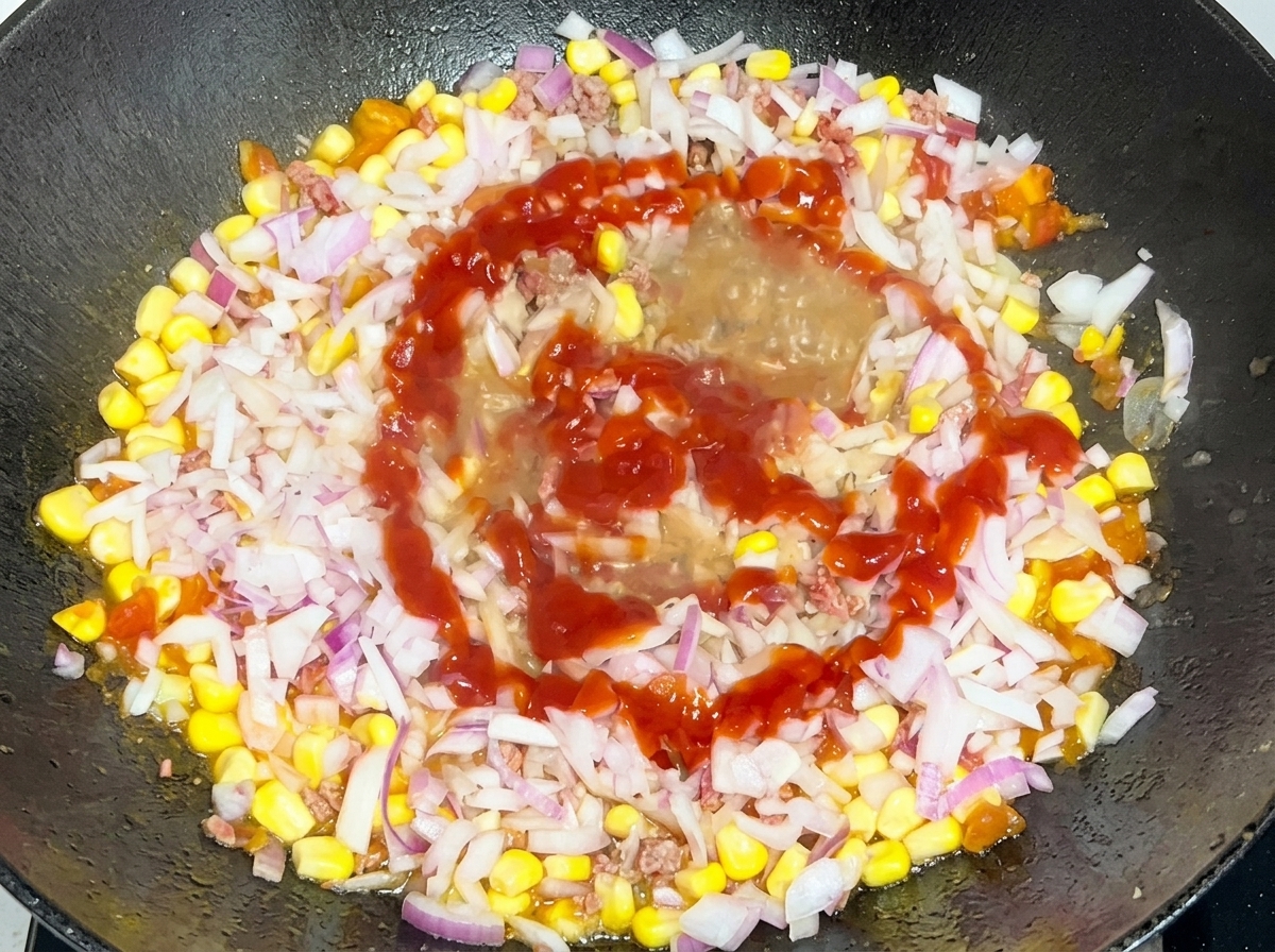 Finely diced red onions and a swirl of red ketchup being added to the beef and corn mixture in the pan.