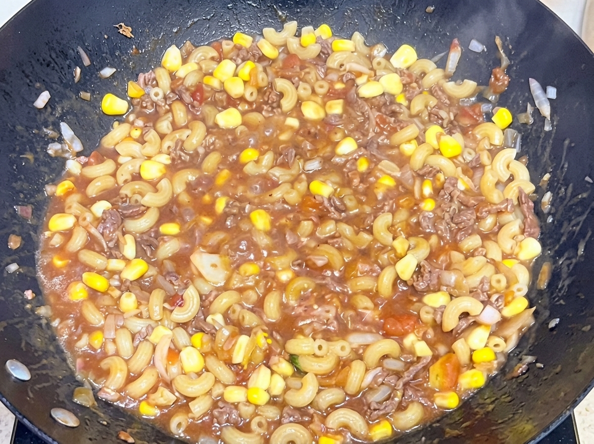 Cooked elbow macaroni being mixed into a savory beef and tomato sauce in a large wok.
