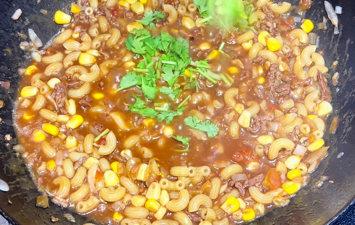 Hand sprinkling chopped green cilantro over a steaming pan of beef and tomato macaroni.