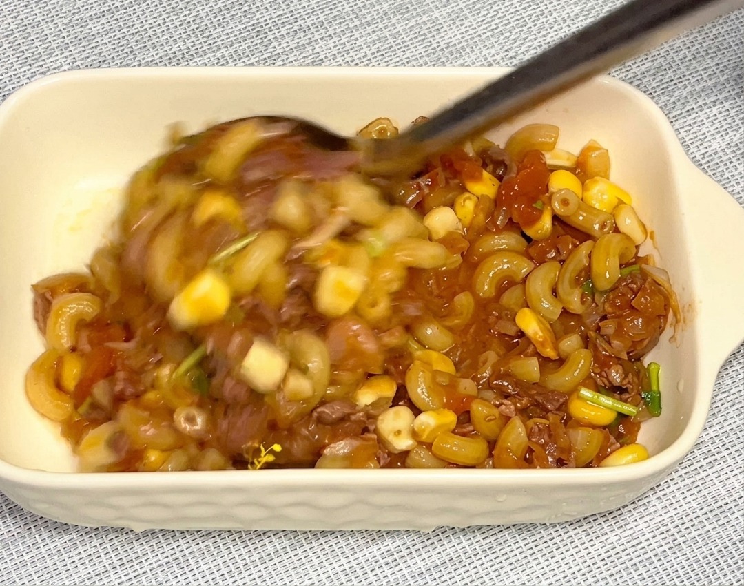 A large metal spoon transferring a mixture of macaroni, corn, and beef into a rectangular white ceramic dish.