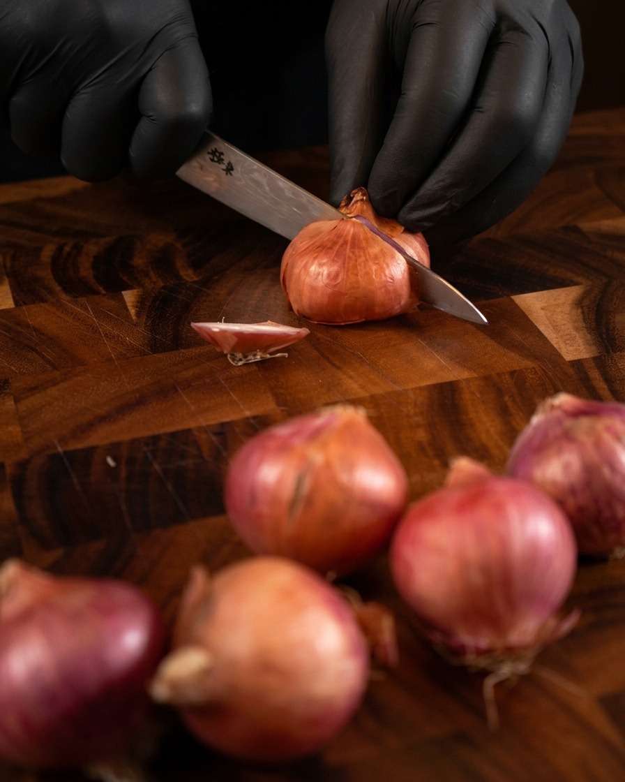 Hands wearing black gloves using a sharp metal knife to thinly slice whole shallots on a dark wood board.