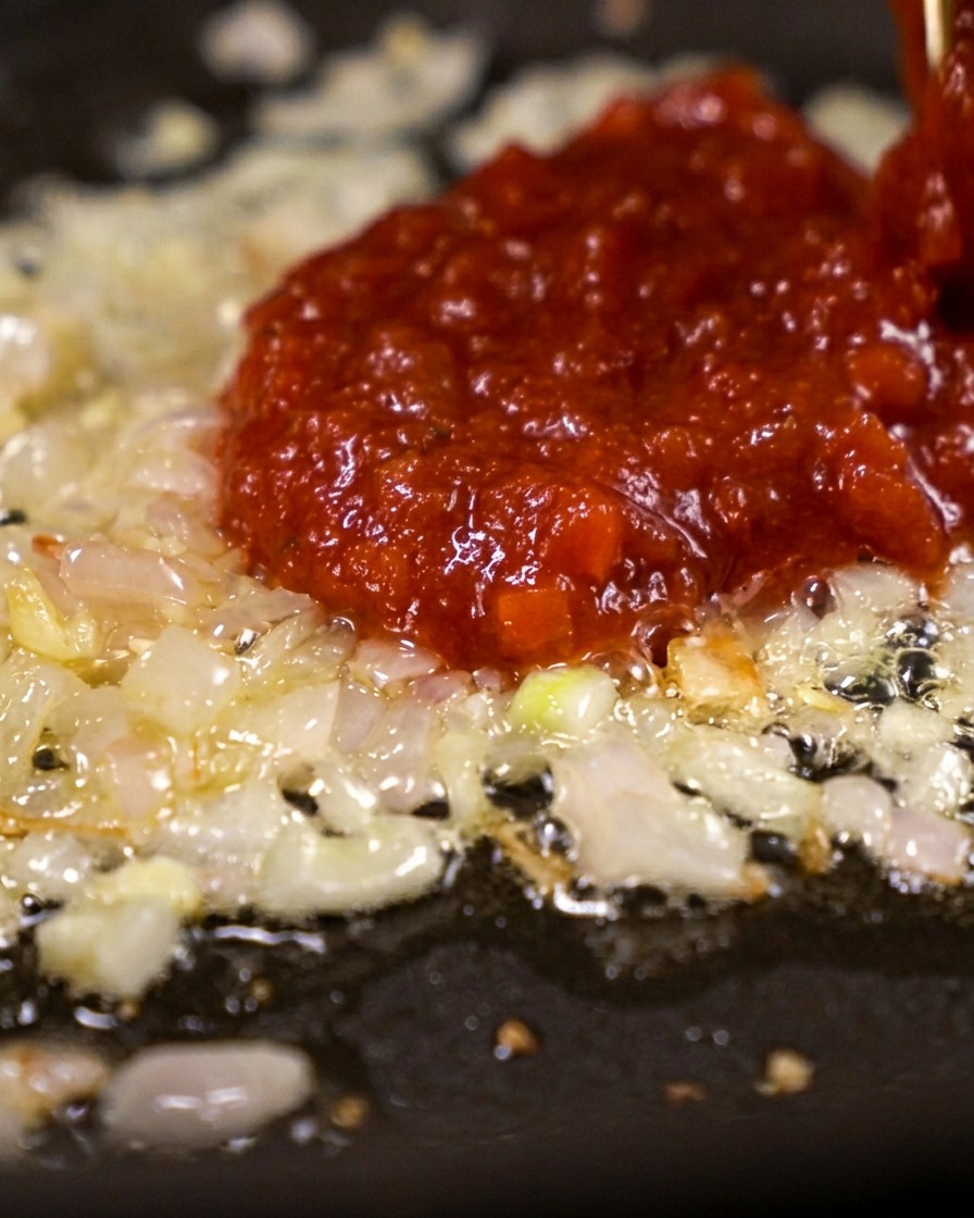 A large spoonful of thick, bright red tomato paste being dropped onto a bed of shimmering sautéed shallots.