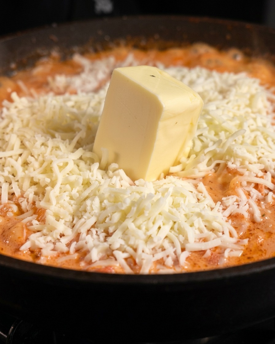 A thick square block of pale butter sitting on top of a pile of shredded white mozzarella cheese over orange pasta sauce.