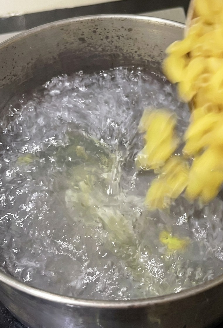 Elbow macaroni being poured from a bowl into a stainless steel pot of bubbling water on a stove.