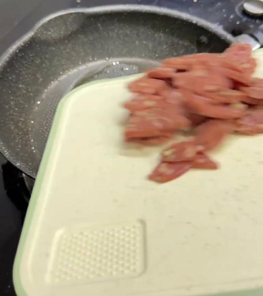 Slices of pink salami falling from a white cutting board into a dark, speckled frying pan.