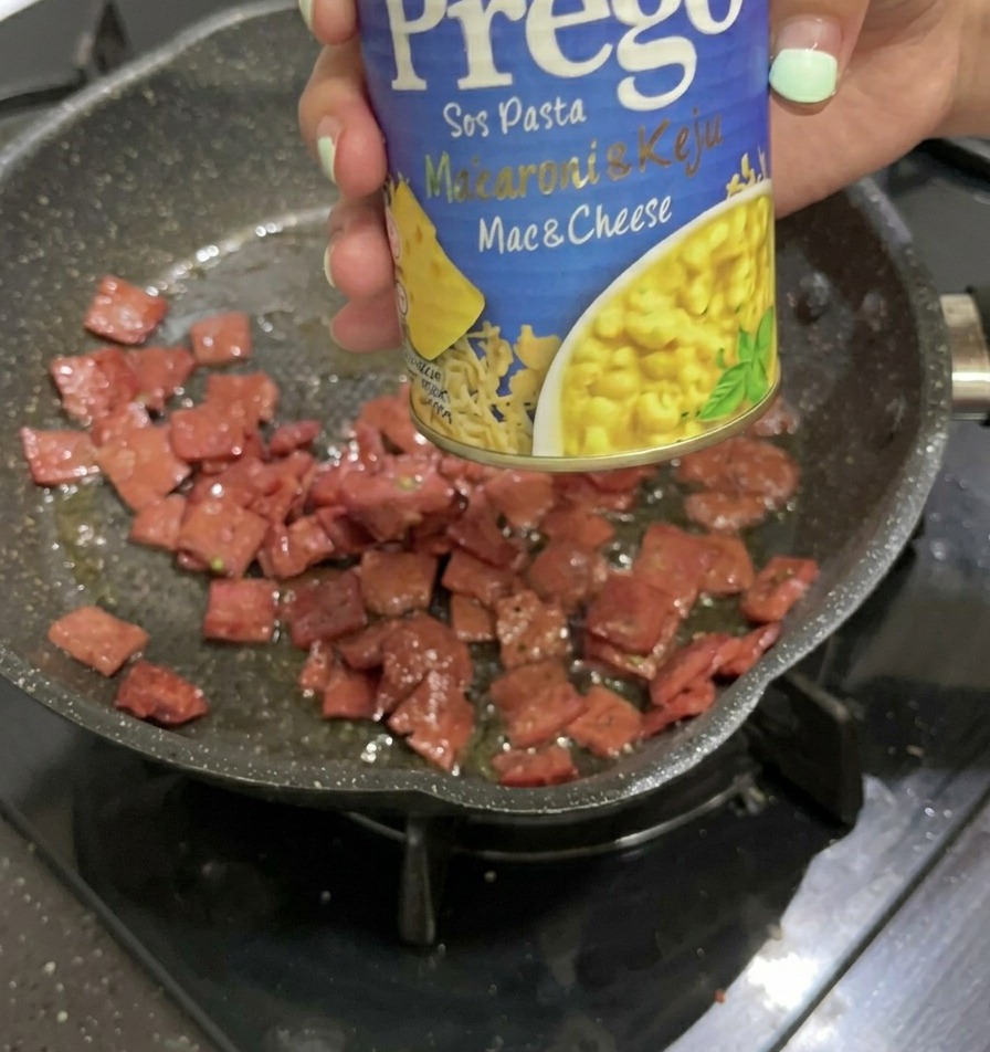 A hand holding a blue and yellow can of Prego Mac & Cheese sauce over a frying pan containing fried salami pieces.
