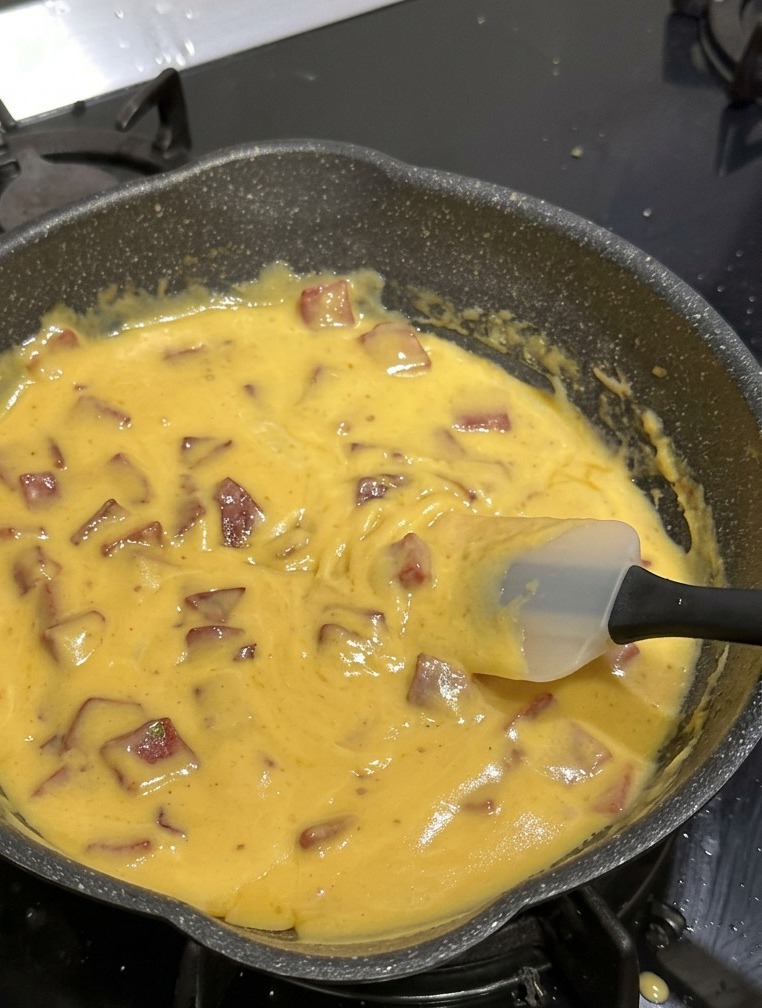 A black non-stick pan filled with elbow macaroni, diced beef salami, and a thick, vibrant yellow cheese sauce being stirred with a white silicone spatula.