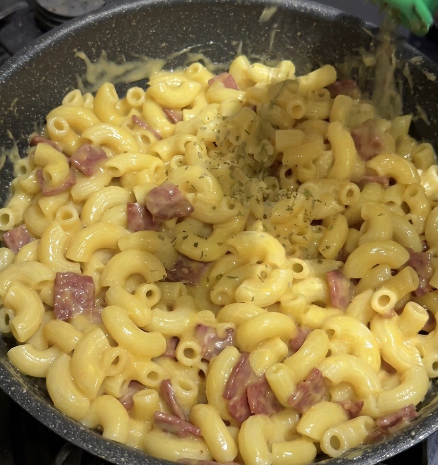 Dried green parsley flakes being shaken from a green spice container onto a pan full of macaroni and cheese.
