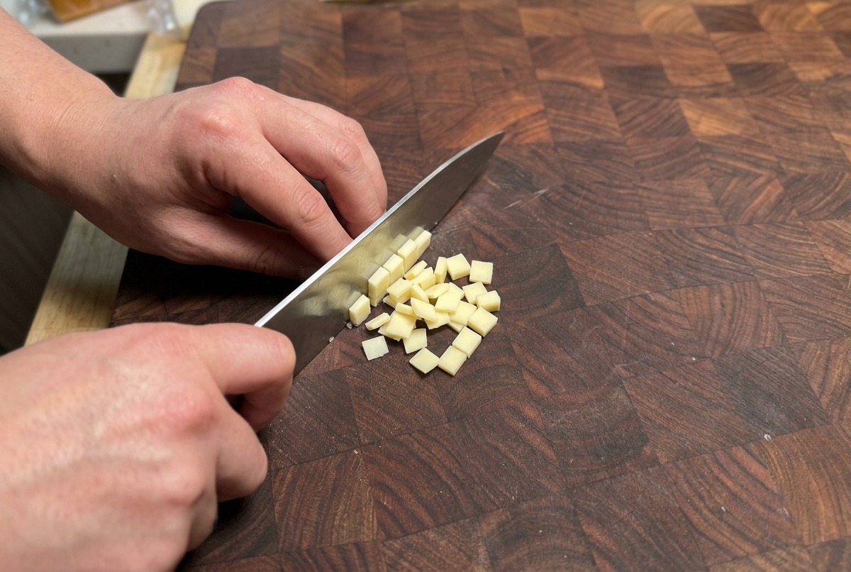 Hands using a chefs knife to dice a block of pale yellow cheese into small cubes on a textured wooden cutting board.