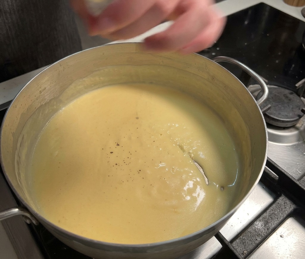 Fresh black pepper being ground into a smooth, thick, creamy yellow cheese sauce simmering in a pot.