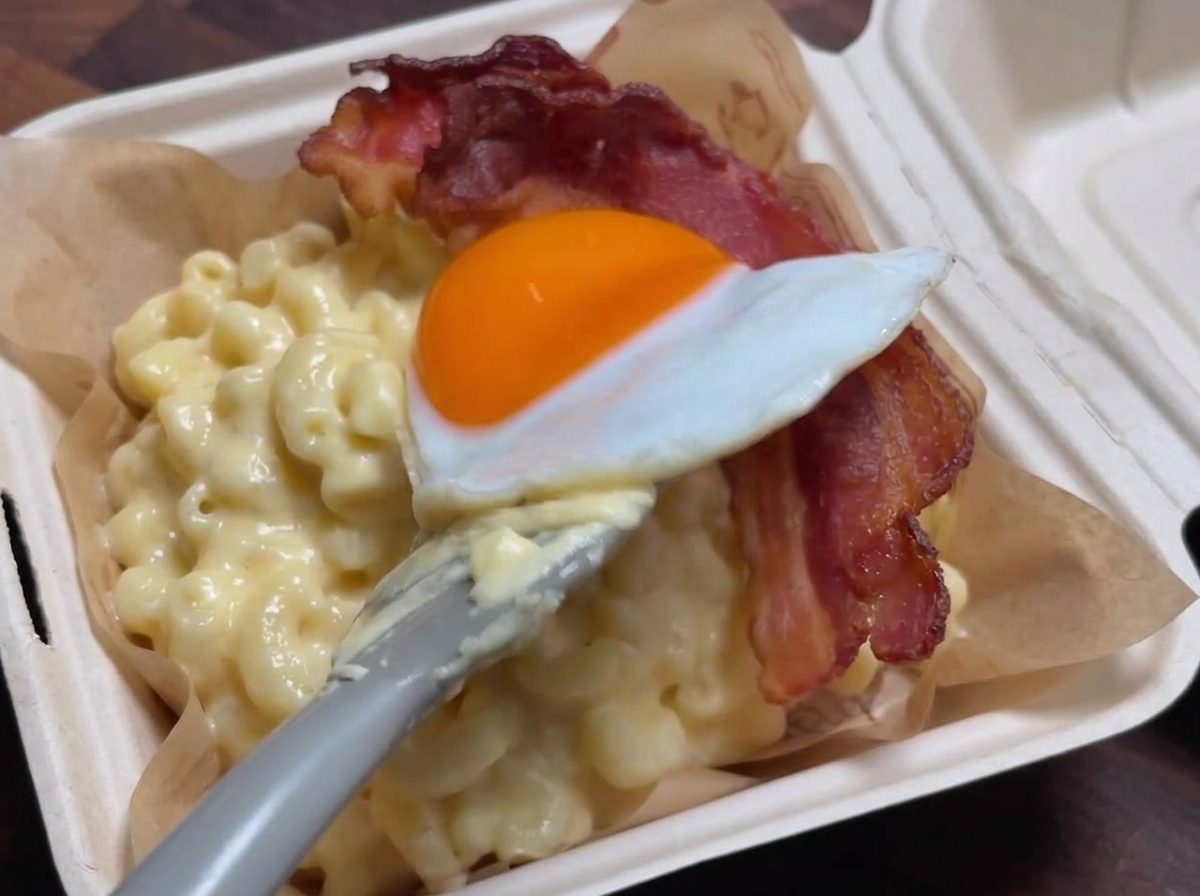 A spatula gently placing a sunny-side-up egg over a strip of crispy bacon resting on top of a serving of creamy macaroni and cheese.