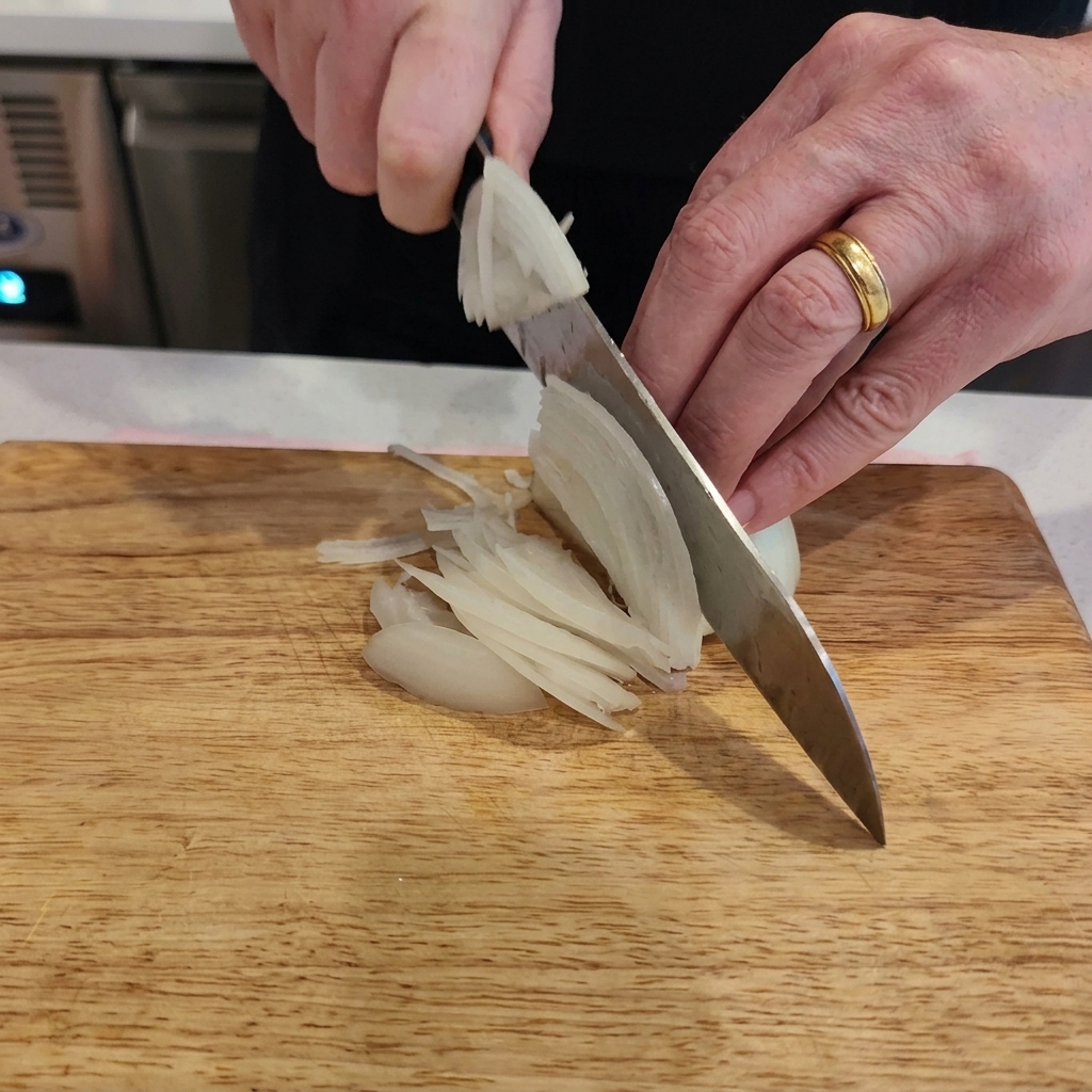 A close-up view of a persons hands using a chefs knife to slice a white onion into thin strips on a light-colored wooden board.