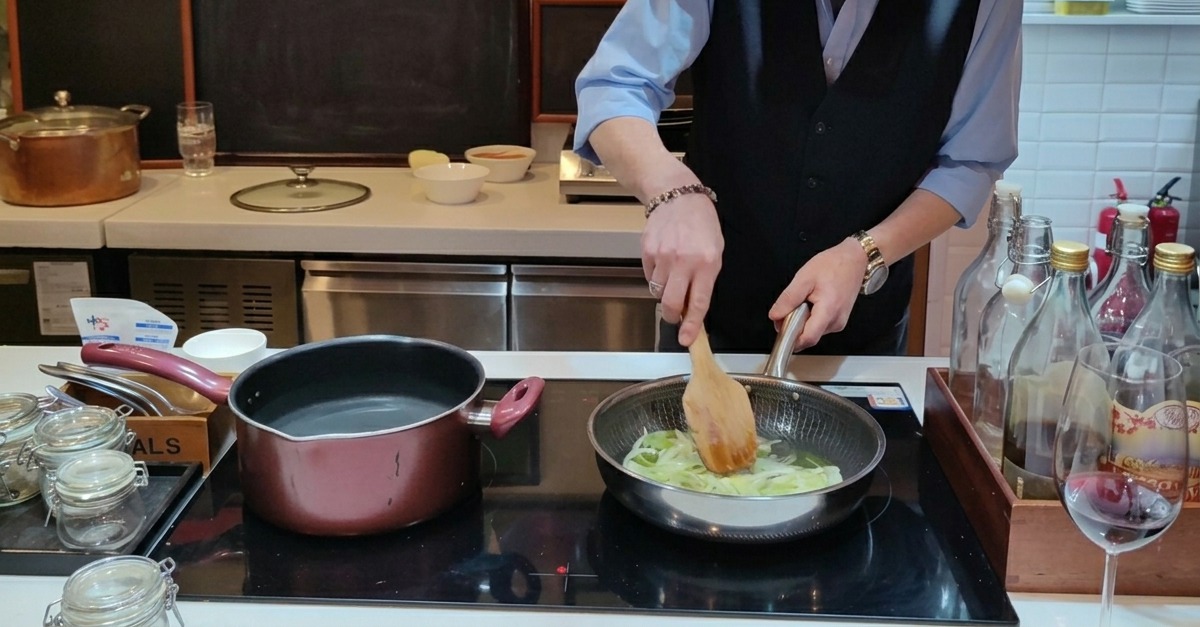 Thinly sliced onions being sauteed in a frying pan with olive oil using a wooden spatula