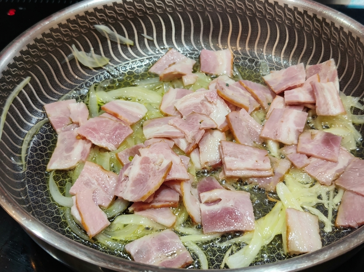 Slices of pink bacon frying in a pan alongside softened, translucent onion strips in oil.