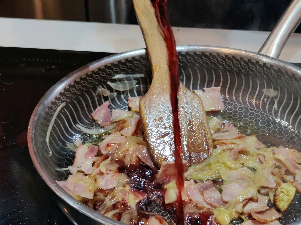 A stream of dark red wine being poured from a glass into a stainless steel pan filled with sizzling bacon and onions.