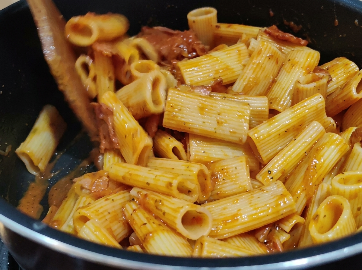Stirring Rigatoni pasta with rich red tomato sauce in a pot using a wooden spatula.